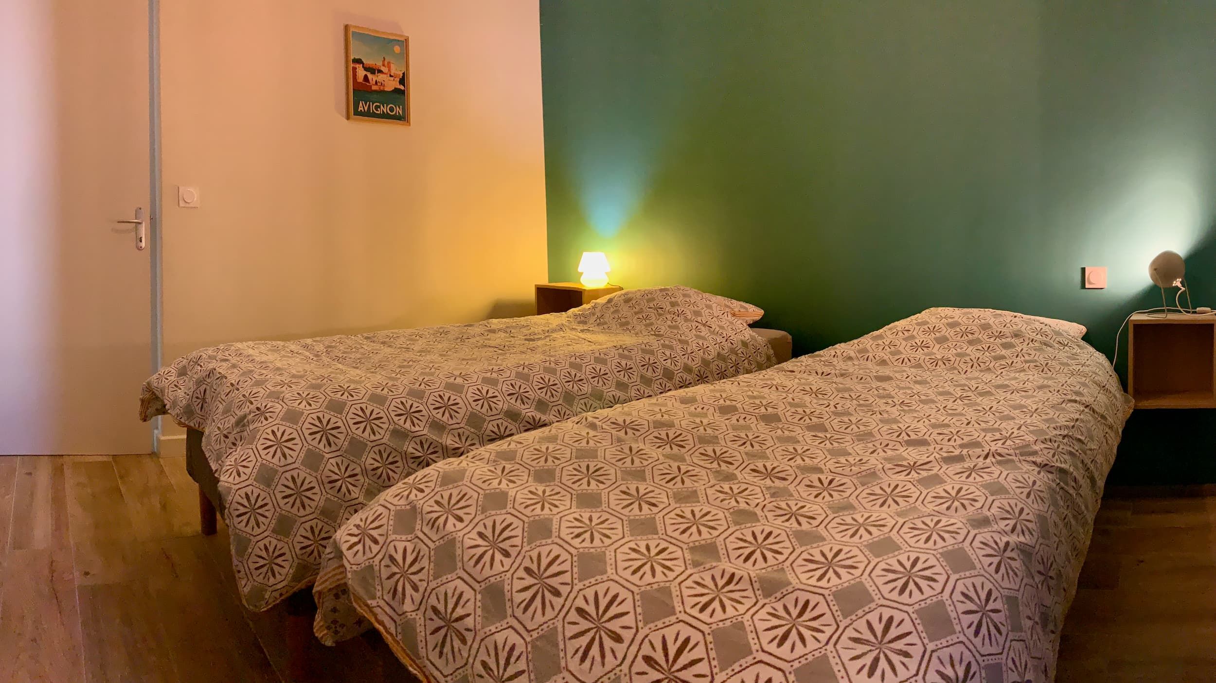 View of the room in the evening with the two beds, teal wall, poster, door, and bedside lamps lit