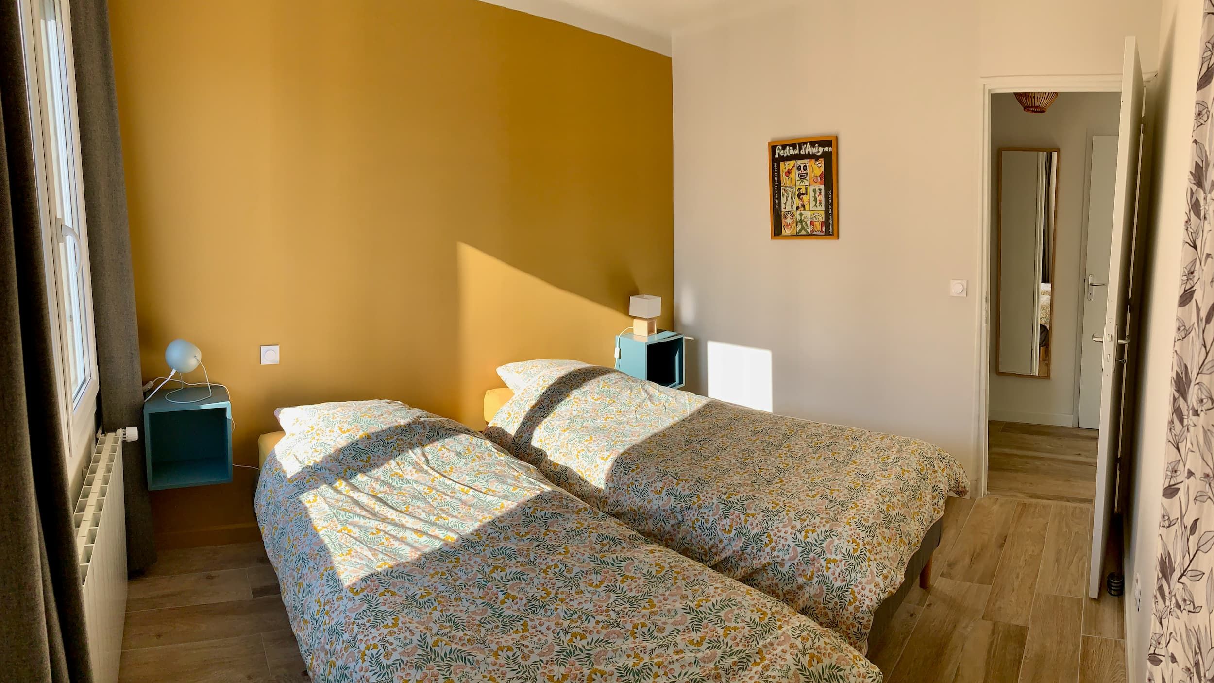 View of the room in the evening with the two beds, the yellow wall, the poster, the door, and the bedside lamps lit